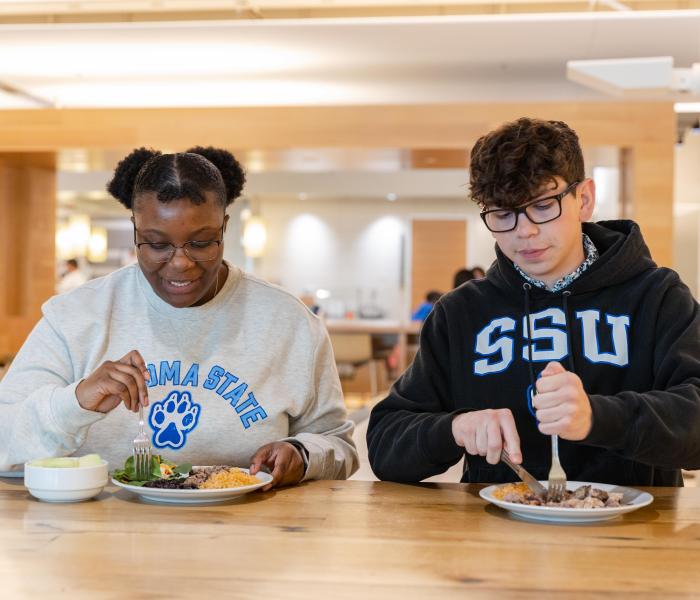 Two students sitting at a table with a plate of food they are starting to cut up to eat
