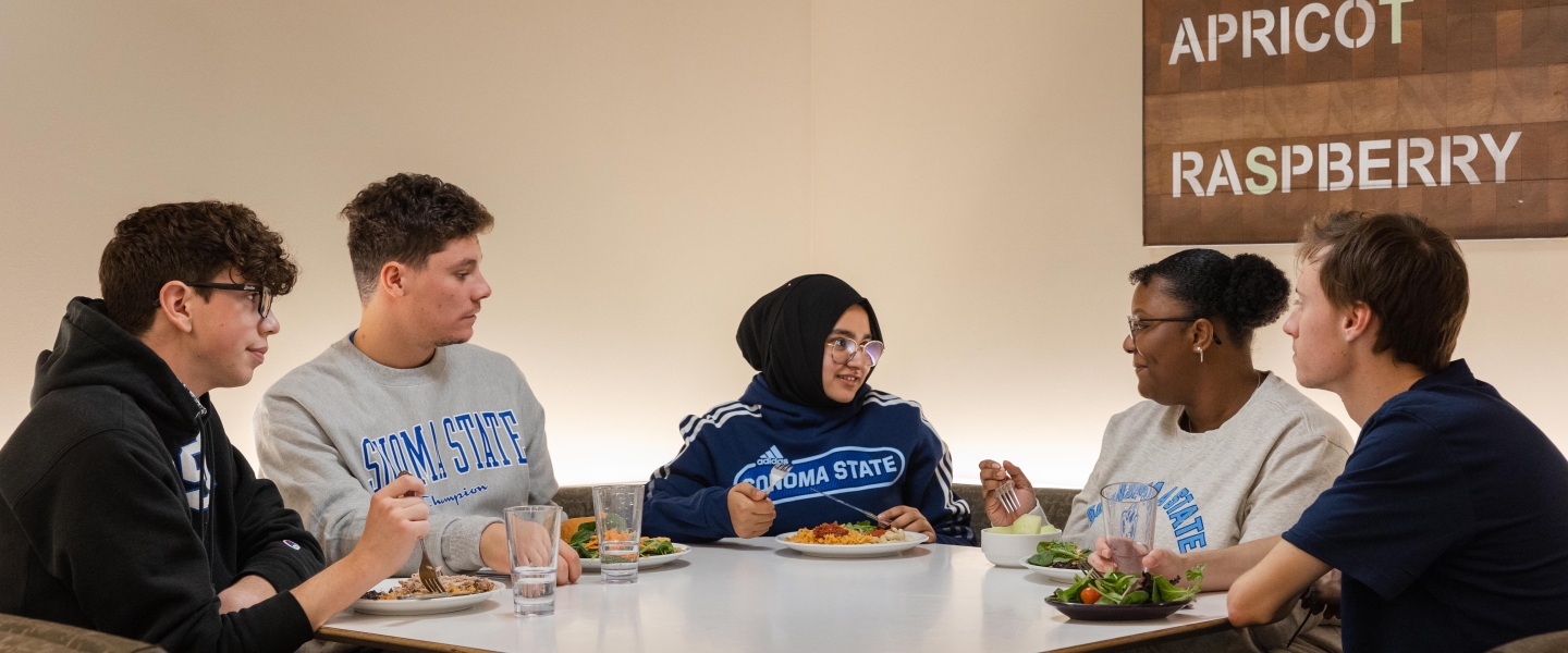 Group of five students sitting at a table talking and eating food