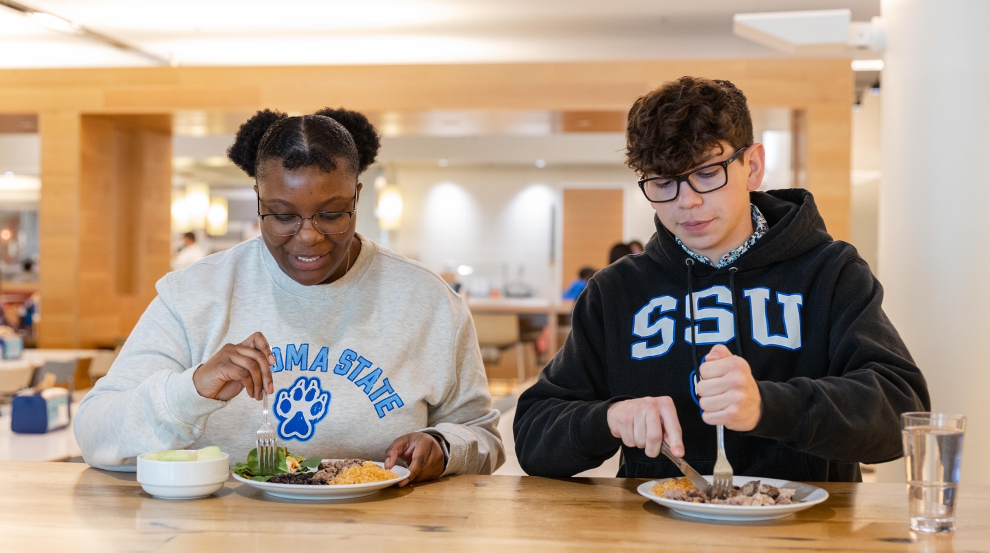 Two students sitting at a table with a plate of food they are starting to cut up to eat