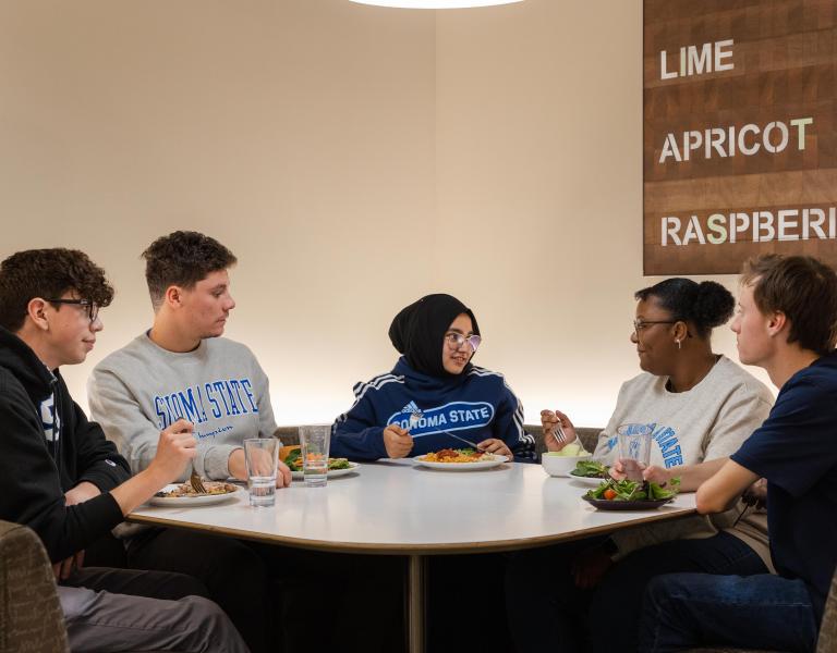 Group of five students sitting at a table talking and eating food