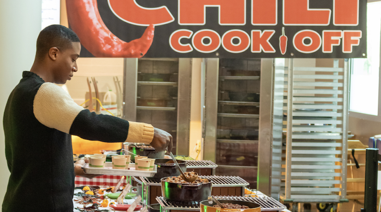 a student serving himself chili from the table with the chili cook off banner in background
