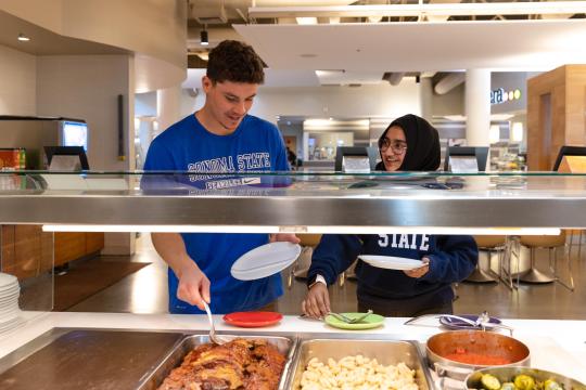 Two students grabbing food from a service area and talking 