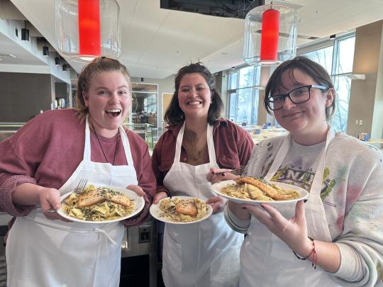 Three students pose with their cooking creation of salmon and pasta on plates