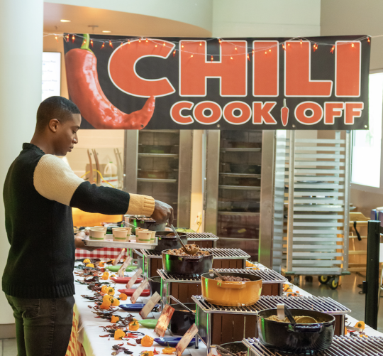 a student serving himself chili from the table with the chili cook off banner in background